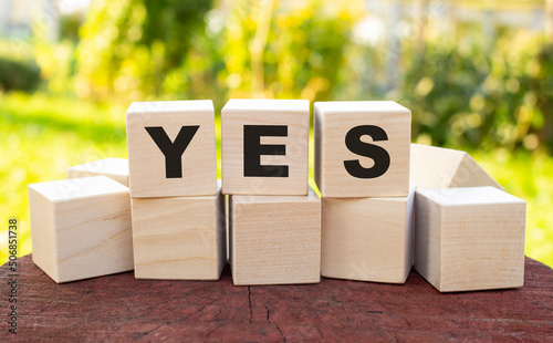 The word YES is made up of wooden cubes lying on an old tree stump against a blurred garden background.
