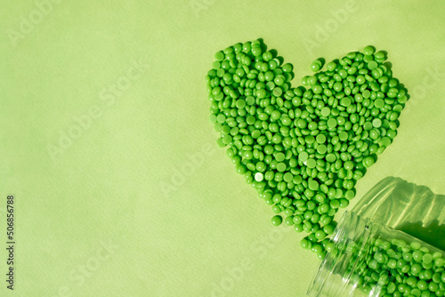 GREEN WAX GRANULES LIE ON A GREEN BACKGROUND, POURED FROM A TRANSPARENT HEART-SHAPED JAR. SPACE FOR TEXT