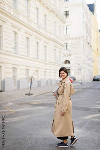happy woman in beige trench coat walking with laptop on street in vienna.