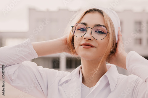 Fototapeta Naklejka Na Ścianę i Meble -  Young female nurse standing outside hospital infirmary. Gorgeous doctor woman dressed white medical gown and cap wear eyeglasses posing, smiling, looking at the camera.
