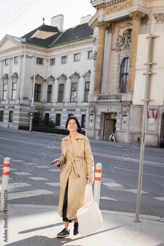 cheerful woman in stylish coat holding bag and smartphone on street in vienna.