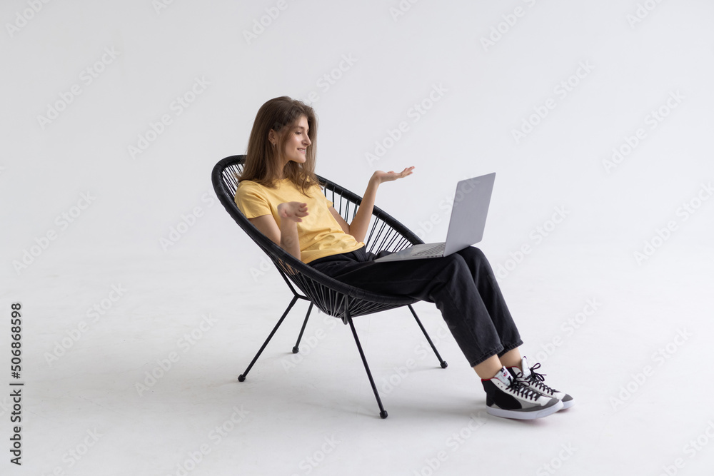 Cheery woman having online video call on laptop, sitting on chair against white wall, full length. Millennial lady participating in webinar, communicating remotely on internet Cheery woman having online video call on laptop, sitting on chair against white wall, full length. Millennial lady participating in webinar, communicating remotely on internet