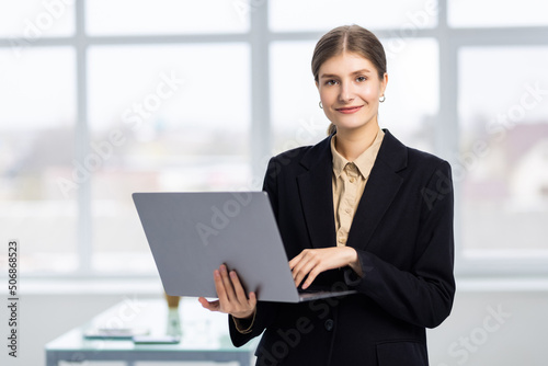 Young confident businesswoman holding laptop computer, standing in office near window. © dianagrytsku