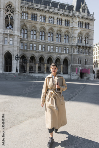 freelancer in trendy beige trench coat walking with laptop on street of vienna.