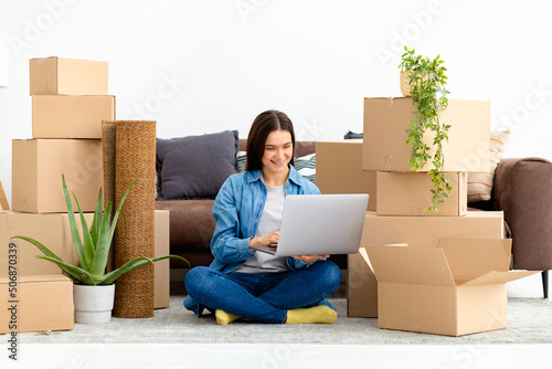 Moving to a new house, rental housing. Happy caucasian woman using laptop computer to search and order transportation service and movers to move to a new home photo
