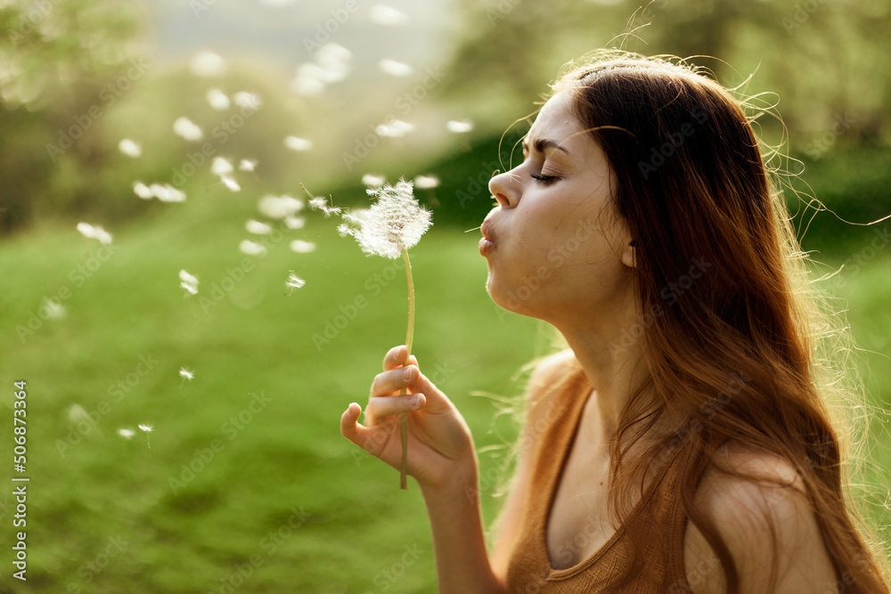 Woman with a dandelion flower in her hands smiling and blowing on it against a background of summer greenery and sunlight Woman with a dandelion flower in her hands smiling and blowing on it against a background of summer greenery and sunlight