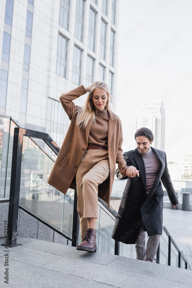 trendy woman in coat holding hands with young man while walking in city.