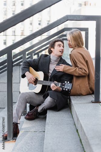 happy blonde woman in autumn clothes touching man playing acoustic guitar on stairs.