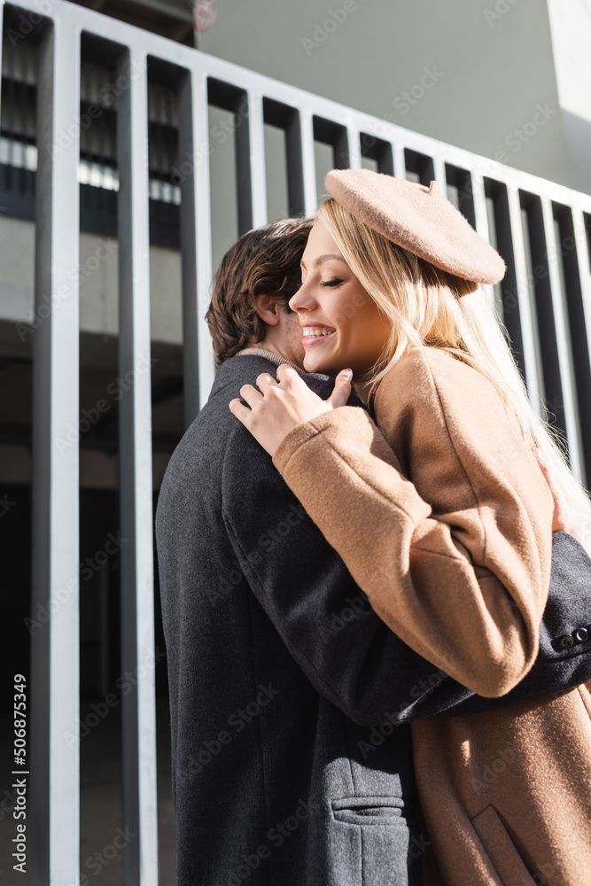 cheerful woman in beret embracing with boyfriend near fence on street. cheerful woman in beret embracing with boyfriend near fence on street.