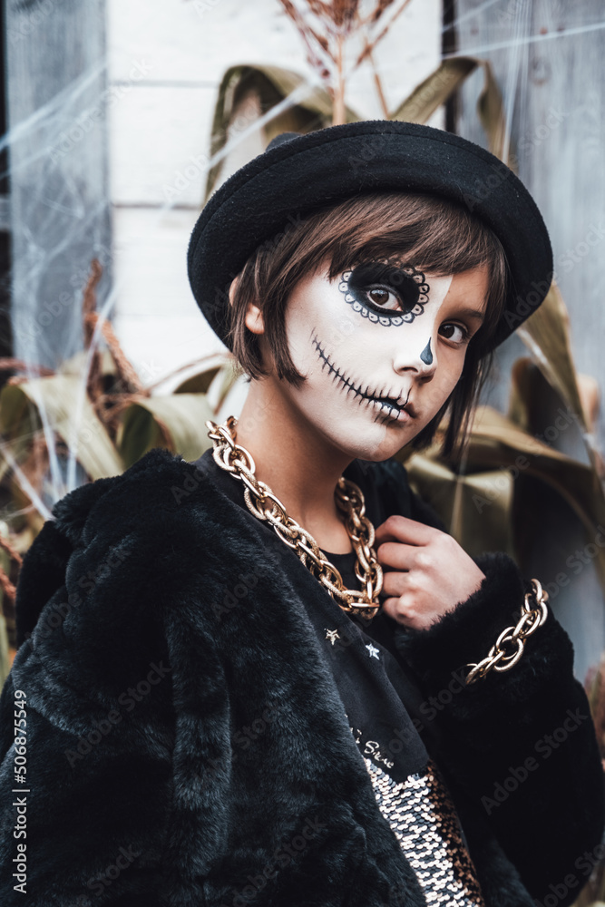 Stockfoto med beskrivningen Beautiful scary little girl celebrating  halloween. Terrifying black, white half-face makeup and witch costume,  stylish image. Horror, fun at children's party in barn on street. Hat, fur  coat, chain