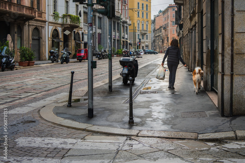 A young woman with a white bag in her hand and a dog on a leash walks through the face of Milan, Italy on a rainy morning, past motorcycles on the side of the road towards the light, real Italy