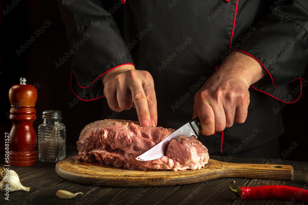 Chef cuts meat with a knife before baking. Spices on the kitchen table for preparing a delicious lunch or dinner