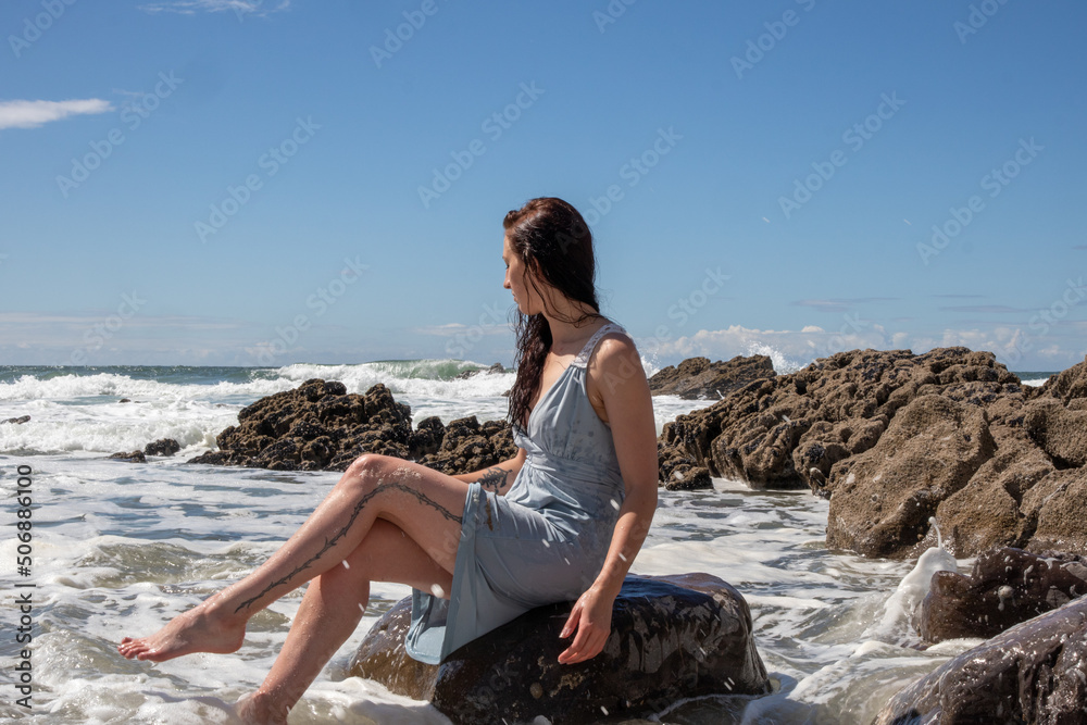 A woman in a blue dress at the beach on a sunny day. The blue sky has some cloud cover. The model sits amongst the rocks as the sea washes waves over her