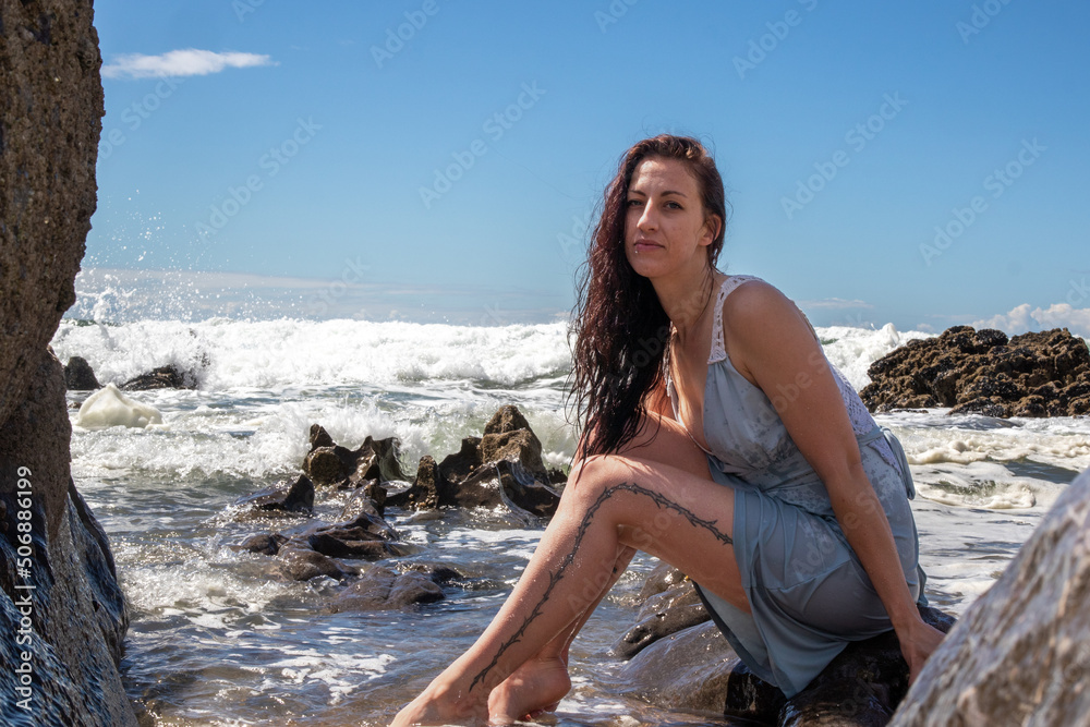 A woman in a blue dress at the beach on a sunny day. The blue sky has some cloud cover. The model sits amongst the rocks as the sea washes waves over her A woman in a blue dress at the beach on a sunny day. The blue sky has some cloud cover. The model sits amongst the rocks as the sea washes waves over her