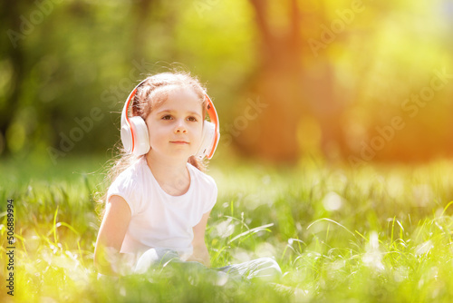 Cute little girl is listening to music in the spring park. Family summer outdoor lifestyle. Happy small kid in headphones sitting on green grass. Beauty nature at summer. Childhood happiness © Dmytro Sunagatov