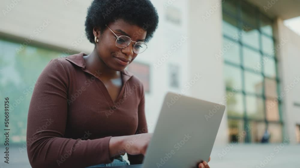 Young female entrepreneur typing email working on laptop outdoors. African American student girl in glasses reading lecture using laptop on the street Young female entrepreneur typing email working on laptop outdoors. African American student girl in glasses reading lecture using laptop on the street