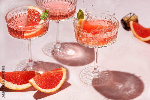 Champagne, rose sparkling wine or paloma cocktails in crystal glasses on pink background with pieces citrus . Refreshing beverage with grapefruit slice and mint. Summer drinks, selective focus photo