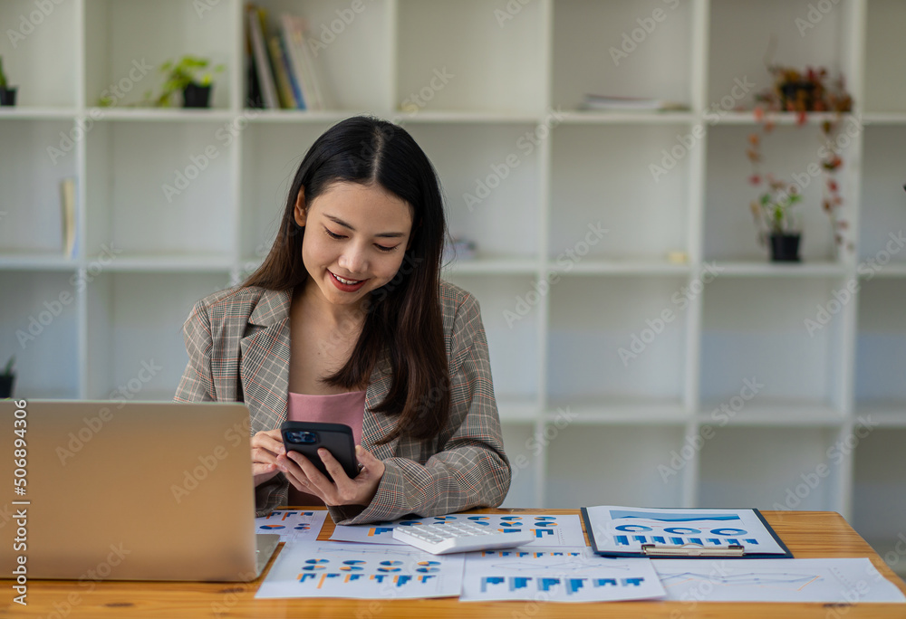 Asian businesswoman holding a smartphone analyzing a report pointing to a graph with a pen, laptop, and calculator. in workstation accountant telemarketing ideas ecommerce free online marketing