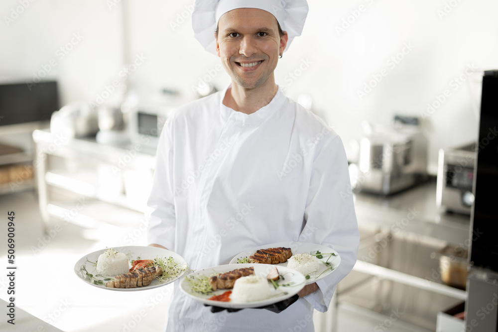 Portrait of young handsome smiling chef in uniform standing with ready meals in the kitchen. Happy caucasian cook working at restaurant. Concept of professional occupation and haute cuisine Portrait of young handsome smiling chef in uniform standing with ready meals in the kitchen. Happy caucasian cook working at restaurant. Concept of professional occupation and haute cuisine