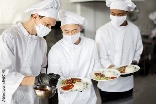 Multiracial team of cooks standing with ready meals for a restaurant in the kitchen. Chefs wearing uniform and face mask during pandemic. Asian chef with Latin and European guys
