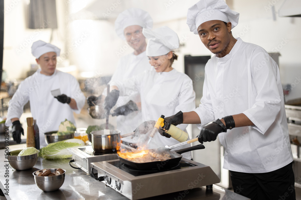Multiracial team of professional cooks in uniform preparing meals for a restaurant in the kitchen. Latin guy burning pan, european cooks making sauce and asian chef managing the process. Teamwork and