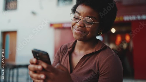 Pretty African girl wearing glasses reading text message on smartphone and smiling. Young woman using mobile phone outdoors