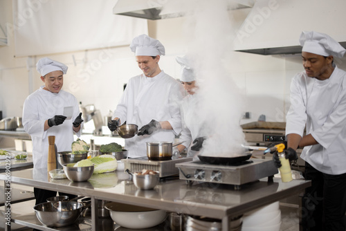 Multiracial team of professional cooks in uniform preparing meals for a restaurant in the kitchen. Latin guy frying meat, european cooks making sauce and asian chef managing the process. Teamwork and