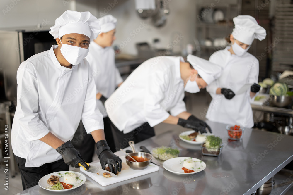 Multiracial group of cooks finishing main courses while working together in the kitchen. Cooks wearing uniform and face mask. Team prepares meals for the restaurant Multiracial group of cooks finishing main courses while working together in the kitchen. Cooks wearing uniform and face mask. Team prepares meals for the restaurant