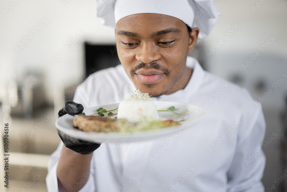 Portrait of young handsome smiling chef in uniform standing with ready meal in the kitchen. Happy Latin-American cook working at restaurant. Concept of professional occupation and haute cuisine Portrait of young handsome smiling chef in uniform standing with ready meal in the kitchen. Happy Latin-American cook working at restaurant. Concept of professional occupation and haute cuisine