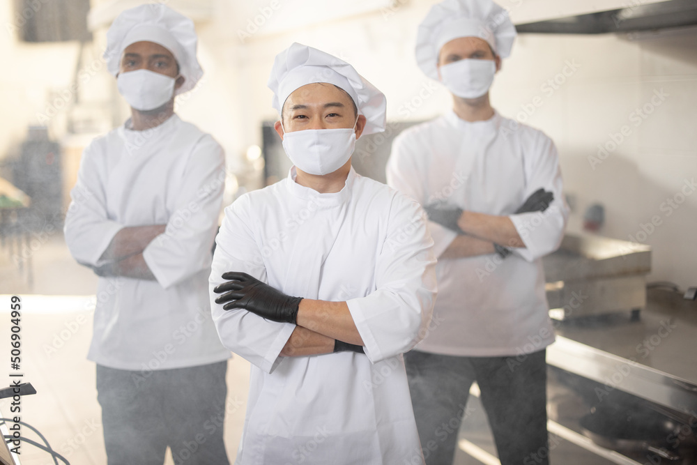 Portrait of three well-dressed chef cooks with different ethnicities standing together in restaurant kitchen. Asian chef standing in front, latin and european guys on background. Cooks wearing face
