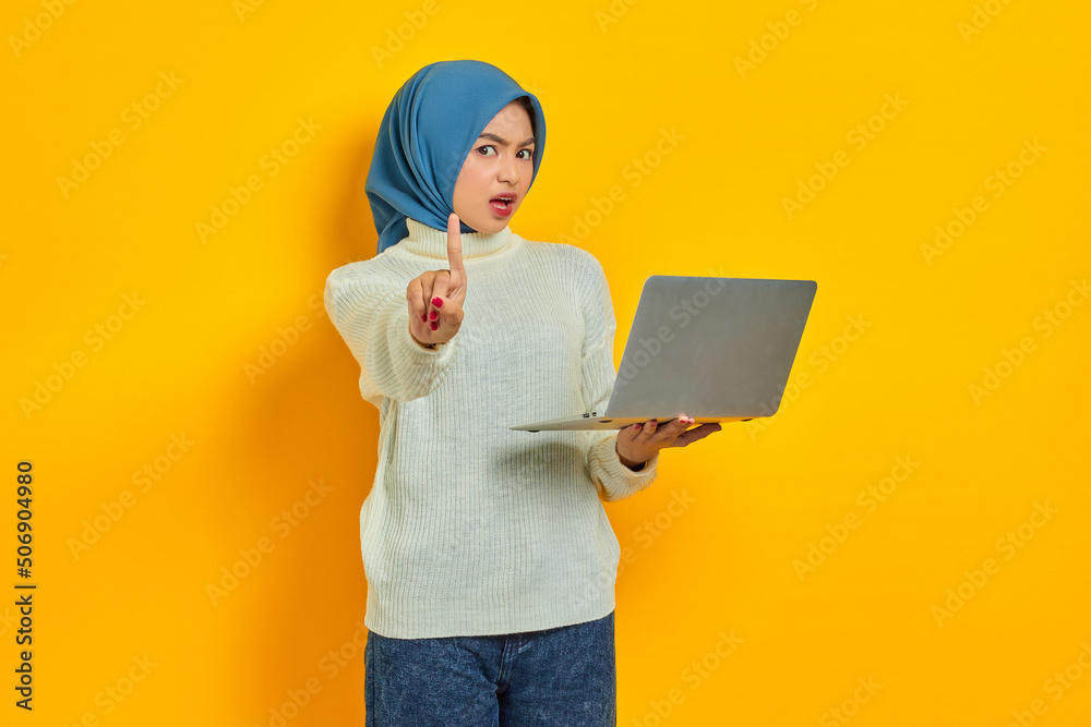 Serious Young Asian muslim woman in white sweater holding laptop and showing stop gesture isolated isolated over yellow background. people religious lifestyle concept Serious Young Asian muslim woman in white sweater holding laptop and showing stop gesture isolated isolated over yellow background. people religious lifestyle concept