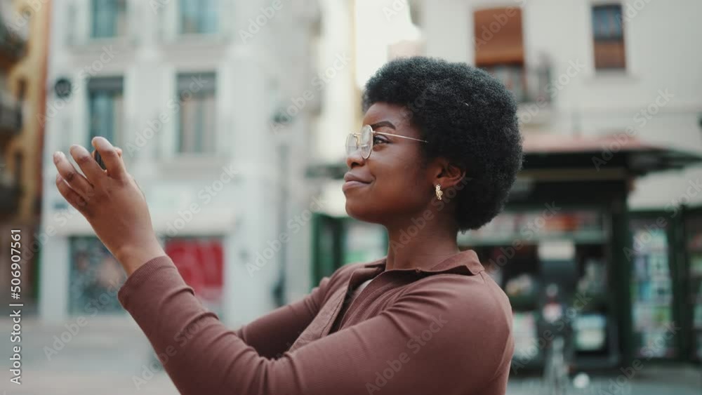 Tracking out side view shot of African American curly girl in glasses taking photo during walk through street. Young woman smiling using smartphone outdoors Tracking out side view shot of African American curly girl in glasses taking photo during walk through street. Young woman smiling using smartphone outdoors