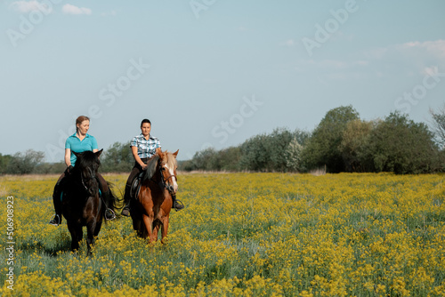 Two women riding horses through blooming field.