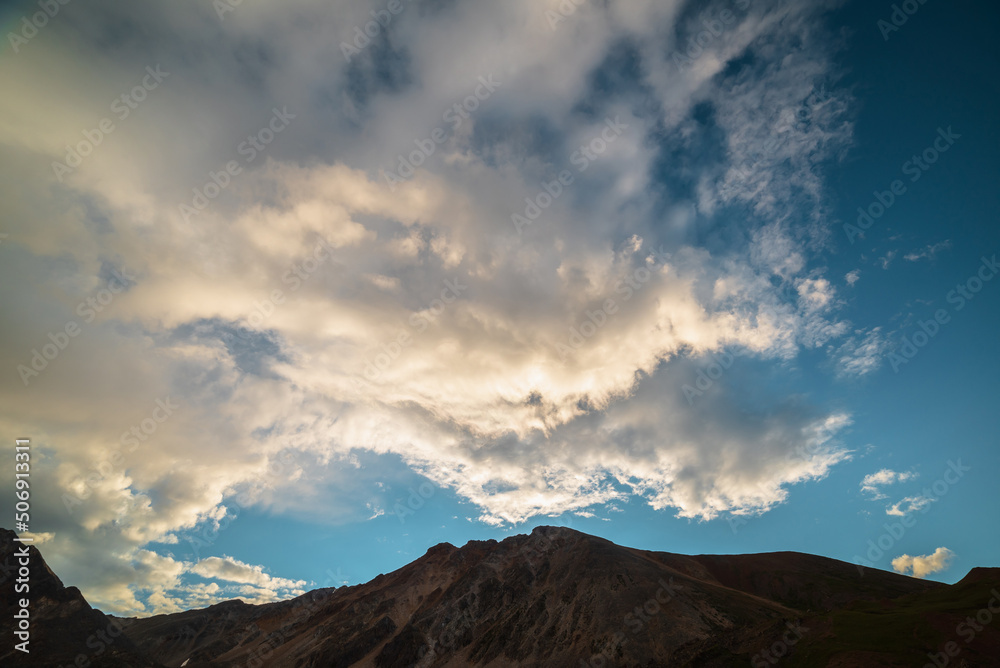 Scenic landscape with dark mountain range silhouette under evening blue sky with orange sunset clouds. Sunlit orange cirrus clouds in sunset sky above silhouette of mountain top at changeable weather.