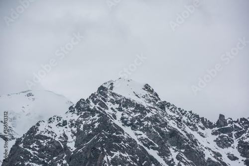 Dramatic landscape with high snowy mountain top in low cloudy sky. White snow on black rocks in low clouds. Awesome view to snow mountain peak in clouds. Monochrome scenery with snowbound mountain top