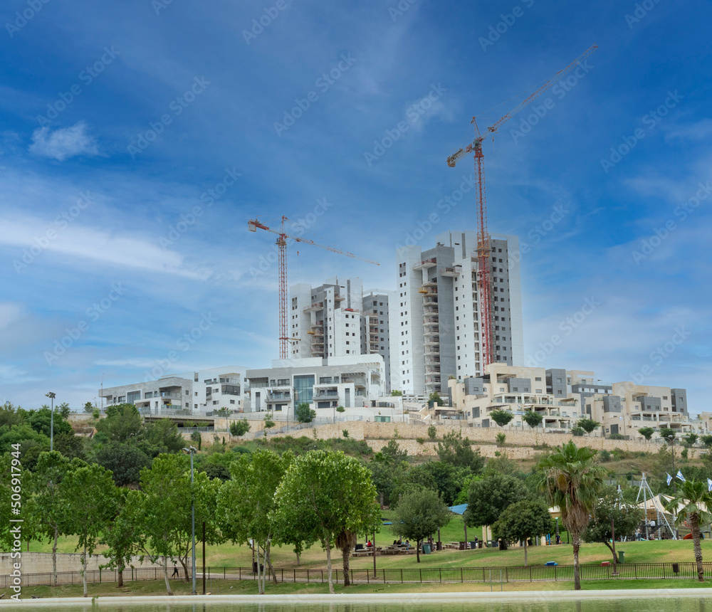 Modiin, Israel, May 21, 2022.Construction of a multi-story residential building overlooking a green park with trees and a lawn next to residential buildings against a blue sky with white clouds. High