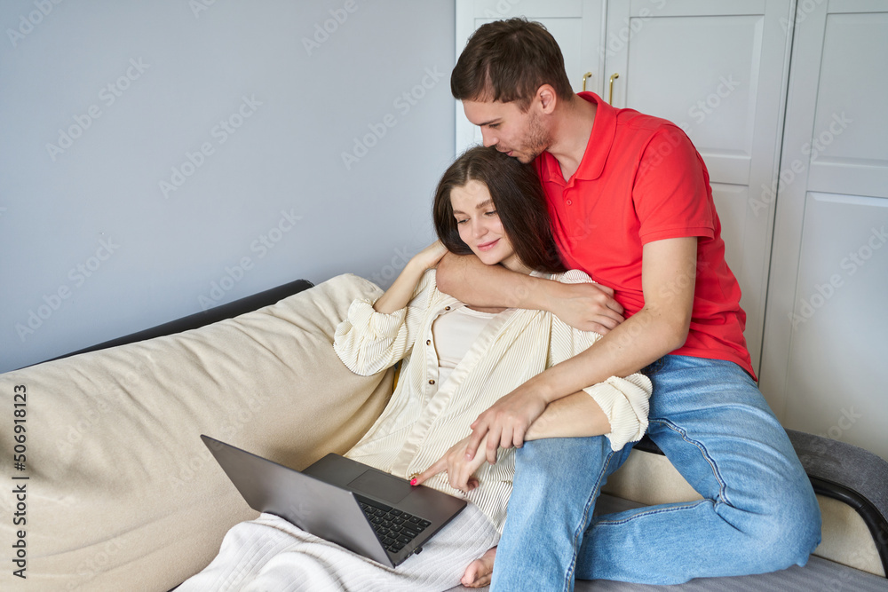 young couple on the sofa. a woman works on a laptop, a man hugs from the back and kisses the top of his head