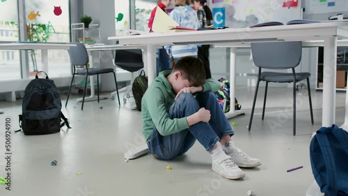 Depressed lonely boy sitting under desk in classroom with classmates playing on background