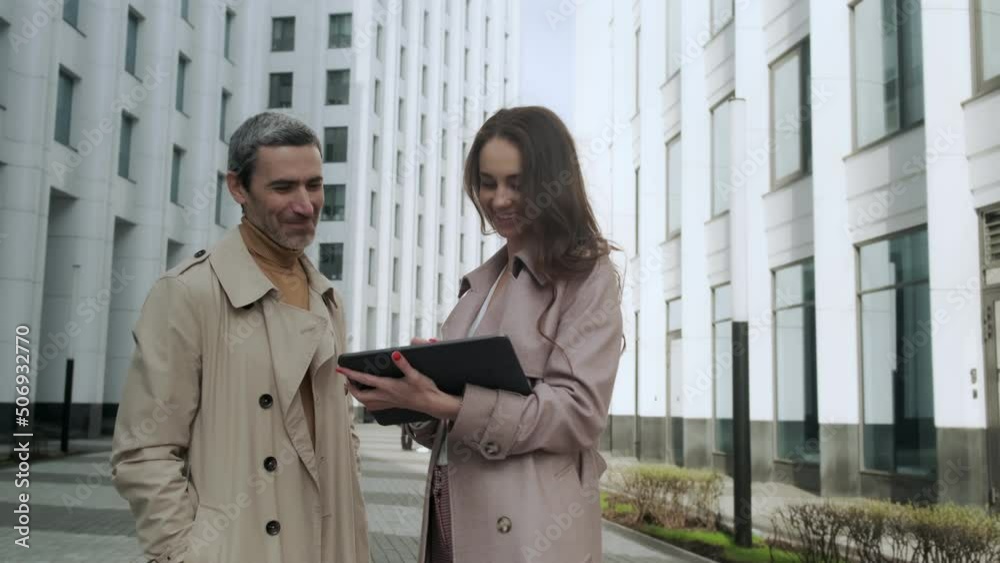 A beautiful girl with a tablet approaches the head of a man standing near office buildings on the street, shows documents on the tablet. The concept of project coordination. A beautiful girl with a tablet approaches the head of a man standing near office buildings on the street, shows documents on the tablet. The concept of project coordination.