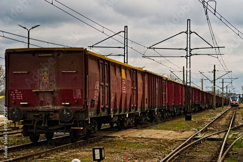 Pociągi ( osobowy i towarowy ) stojące na sąsiednich torach przy peronie na dworcu kolejowym . Widoczne słupy i kable elektrycznej trakcji zasilającej pociągi elektryczne .