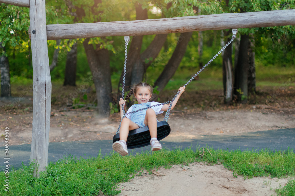 A child plays on an outdoor playground on a summer day. An active happy little girl is riding on a swing. Healthy summer activity for children in warm weather. The little cute baby is rocking. A child plays on an outdoor playground on a summer day. An active happy little girl is riding on a swing. Healthy summer activity for children in warm weather. The little cute baby is rocking.
