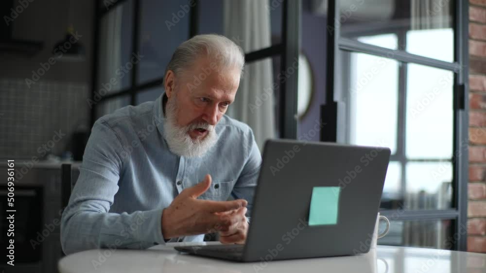 Angry gray-haired mature elderly man having emotionally unpleasant conversation via video call on laptop with family, friends, boss, employees, doctor sitting at kitchen table. Shooting in slow motion Angry gray-haired mature elderly man having emotionally unpleasant conversation via video call on laptop with family, friends, boss, employees, doctor sitting at kitchen table. Shooting in slow motion