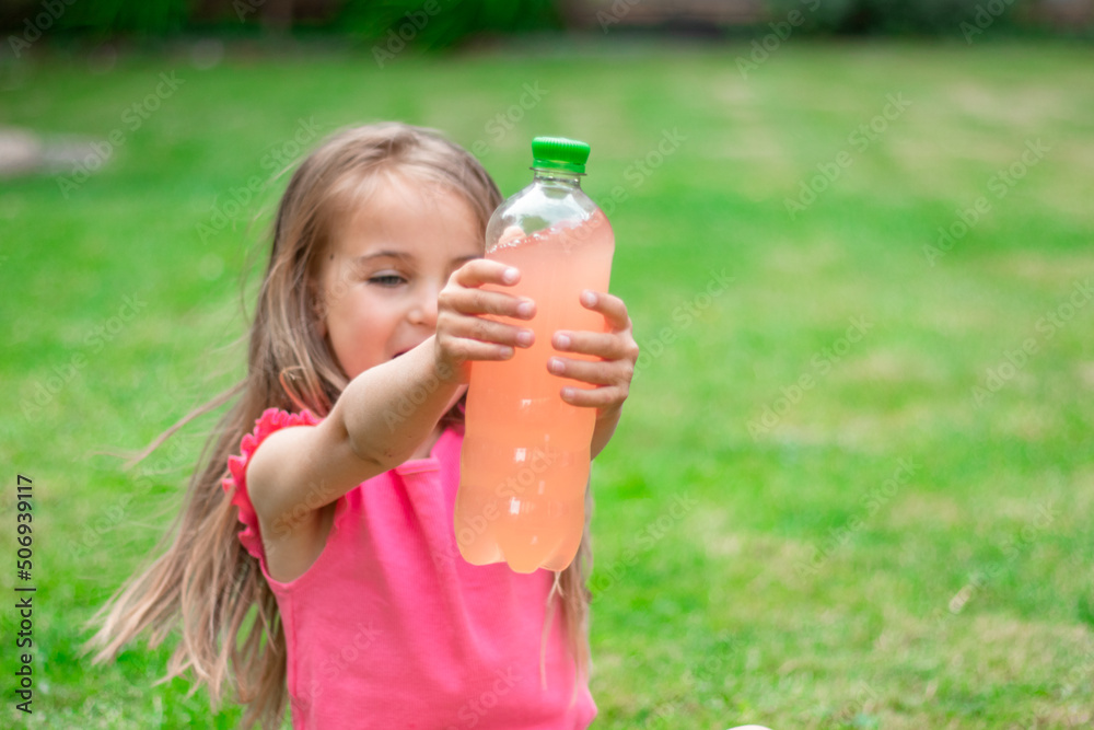 Drink with electrolytes in a transparent bottle in the hands of a happy child on a green lawn in a pink T-shirt Drink with electrolytes in a transparent bottle in the hands of a happy child on a green lawn in a pink T-shirt