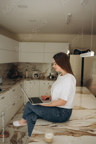 Caucasian female student on video call with friends typing on laptop sipping hot coffee working in kitchen