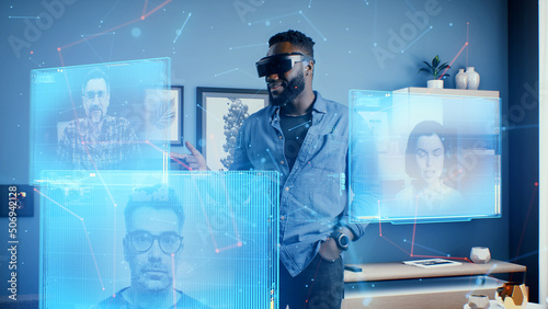 Afro-american man wearing VR goggles at online business meeting in meta universe cyberspace talking with colleagues over video conference, holographic windows in front of him
