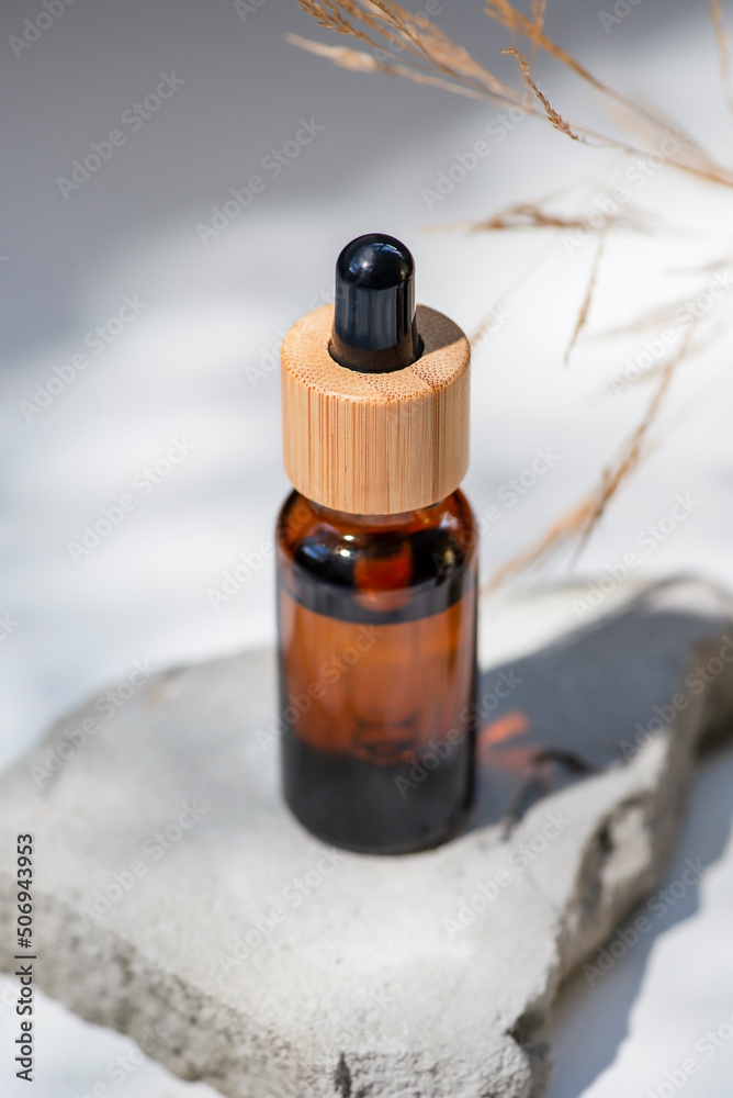 Amber glass dropper with bamboo lid and dry plant. Bottle on grey concrete podium for product presentation. Skincare cosmetic with wildflowers. Beauty concept