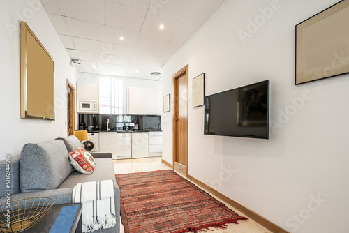 Studio apartment with a furnished living room and an open kitchen with white furniture, a TV hanging on the wall and a red jarapa on the marble floor photo