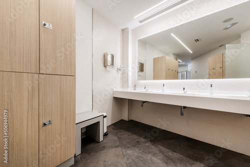 Restroom of a gym locker room with a wooden locker cabinet and a white resin washbasin with a mirror integrated into the wall