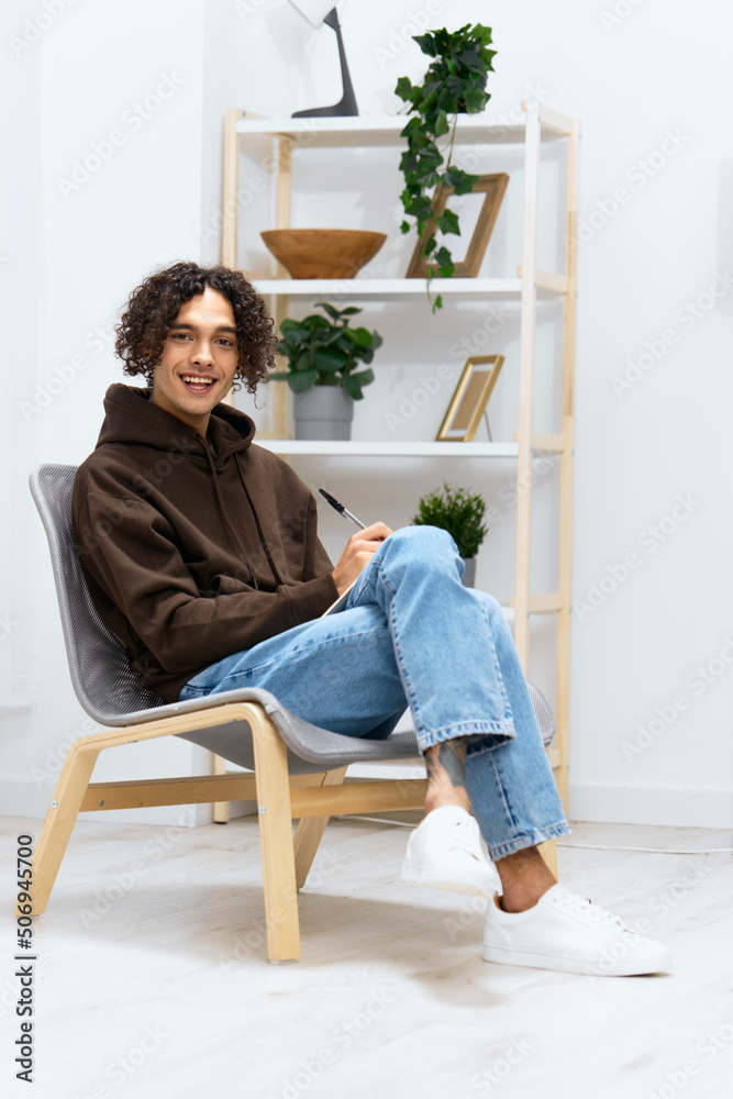 guy with curly hair sit on a chair with a notepad and a writing pen light background guy with curly hair sit on a chair with a notepad and a writing pen light background