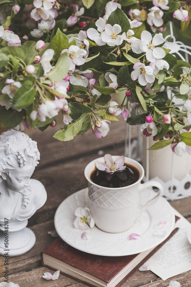 A beautiful postcard. A white coffee cup with a saucer, a statuette, candles, a book and a vase with a bouquet of blooming apple trees. Beautiful still life. Spring time. The concept of A beautiful postcard. A white coffee cup with a saucer, a statuette, candles, a book and a vase with a bouquet of blooming apple trees. Beautiful still life. Spring time. The concept of